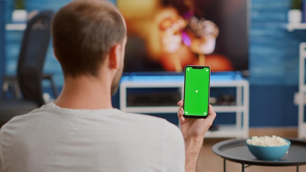 Photo closeup of man holding vertical smartphone with green screen in online conference or group video call in home living room. person using touchscreen mobile phone with chroma key watching webinar.