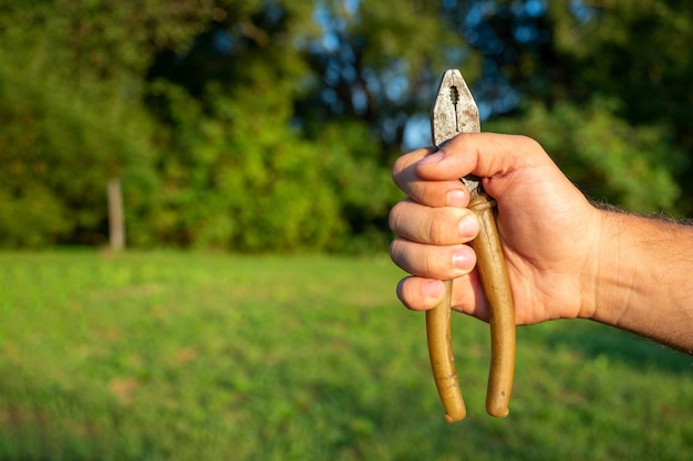 Photo closeup of a man's hand holding old dirty pliers against a green landscape sunlight tools copy space