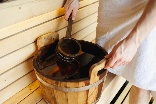Closeup of a man in a sauna scooping water with a ladle from a wooden bucket