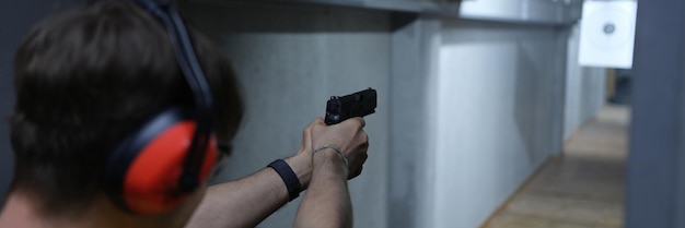 Closeup of man shooter in tactical headphones aiming pistol at target in shooting range