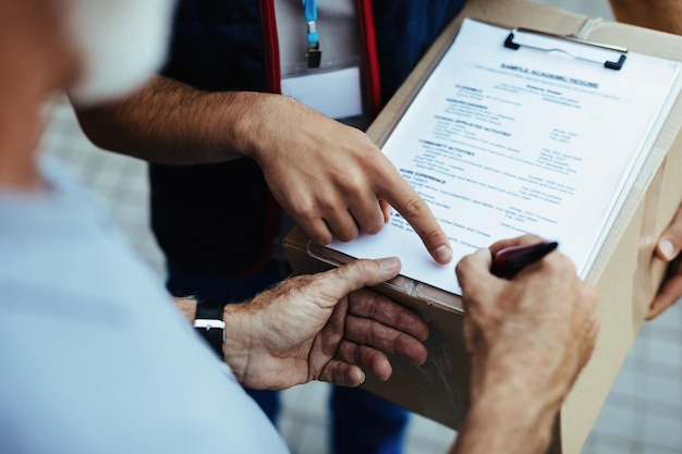 Closeup of man signing paperwork while courier is pointing at place he needs to sign for package delivery