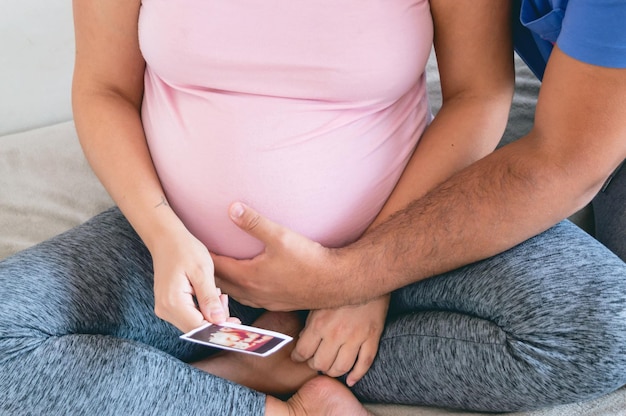 closeup man touches belly and pregnant wife holds ultrasound scan sitting on bed