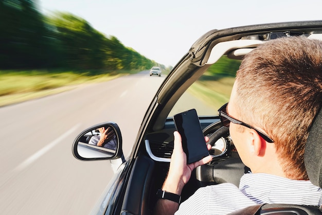 車を運転中にスマートフォンを使用して男のクローズアップ。対向車に向かって運転します。危険な動き。電話で気が散る。男は運転中に電話でメッセージを書きます。ぼやけた動きに影響を与える