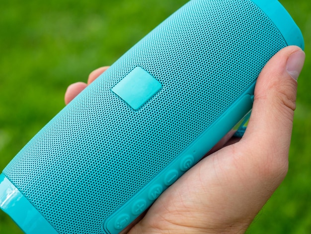 Photo closeup of a mans hand holding a blue wireless music speaker green blurred background