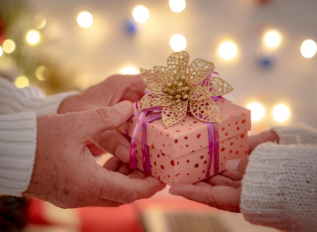 Closeup on mature hands exchanging a christmas present senior\
man offering a gift to an elderly woman lights on backhground