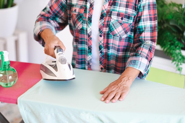 Closeup of mature woman in checkered shirt ironing clothes at home on ironing board