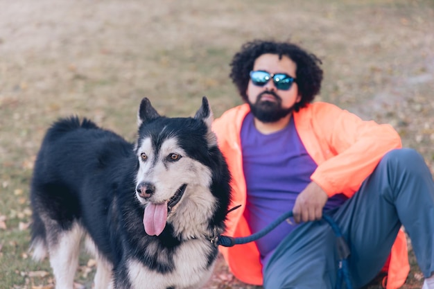 Closeup of a Mexican man with curly hair beard and sunglasses at a park with his husky dog