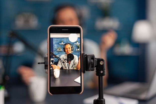 closeup of mobile phone recording smiling woman vlogger on live stream podcast. influencer using microphone and headphones, talking in front of phone camera and filming vlog for social media channel.