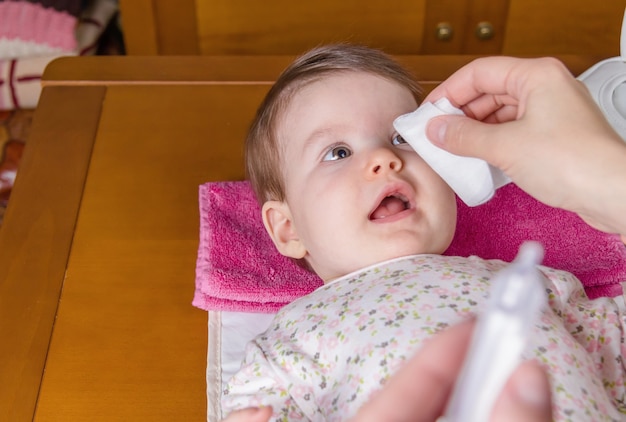 綿の生理的血清で赤ちゃんの目を掃除する母の手のクローズアップ