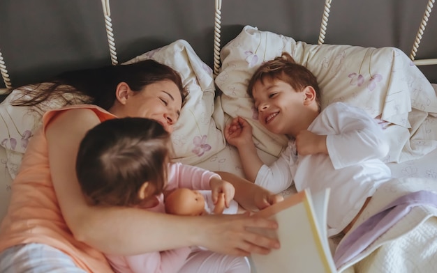 Closeup of mother reading story book to her daughter and son kids lying in the bed. Weekend family leisure time concept.
