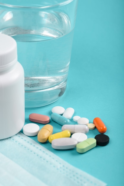 Разноцветные таблетки крупным планом со стаканом воды, медицинская маска на синем фоне, концепция профилактики