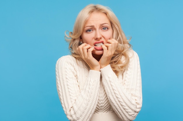 Closeup nerveus angstig blonde vrouw in witte trui haar nagels bijten op vingers in paniek wachten op testresultaten fobie Indoor studio opname geïsoleerd op blauwe achtergrond