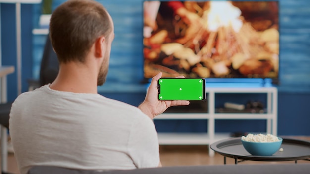 テレビの前のソファに座ってオンラインビデオコンテンツを見ている緑色の画面でスマートフォンを保持している男のクローズアップ。インフルエンサーvlogを楽しんでいるクロマキー付きのタッチスクリーン携帯電話を見ている人。