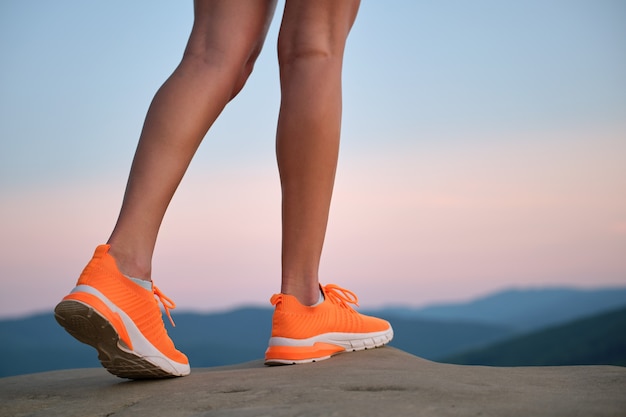 夏に山のハイキングコースを歩いている明るいオレンジ色のスニーカーの靴で若い女性のスリムな脚のクローズアップ。新鮮な空気の概念に関するアクティブな生き方と運動。