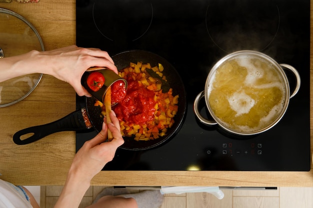 소스를 준비하는 동안 껍질을 벗긴 토마토 통조림을 프라이팬에 추가하는 여성 손의 클로즈업 오버헤드 보기