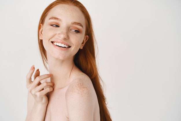 Closeup of pale redhead woman standing naked on white background turn right and smiling at copy space about skincare