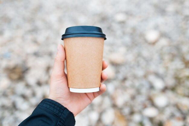 Closeup of paper cup for coffee takeaway in male hand on background of blurred rocky ground