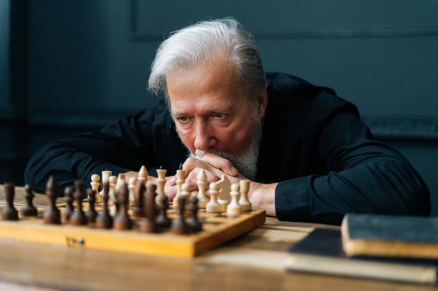 Closeup of pensive grayhaired senior older man thinking game strategy sitting on wooden table with chess board