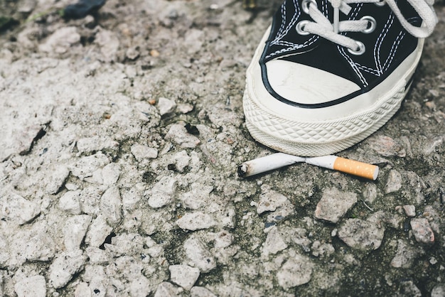 Photo closeup of a persons foot wearing black sneakers shoes crushing a cigarette butt on asphalt