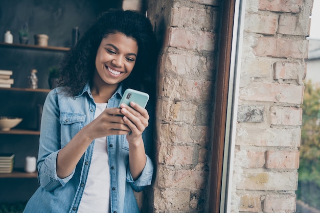 Closeup photo of amazing dark skin curly lady holding telephone reading boyfriend sms leaning brick window wall wear casual denim outfit modern flat indoors