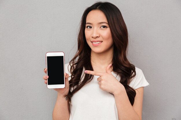 Closeup photo of asian woman in casual t-shirt demonstrating copyspace screen of modern smartphone, isolated over gray wall