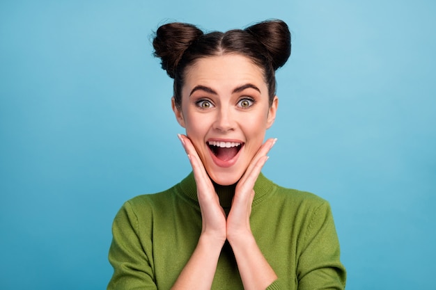 Closeup photo of attractive crazy lady two funny buns good mood open mouth arms on cheeks wear warm green turtleneck pullover isolated blue color wall