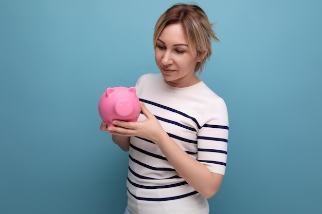 Closeup photo of blond charming young woman in casual outfit thoughtfully holding a piggy bank with