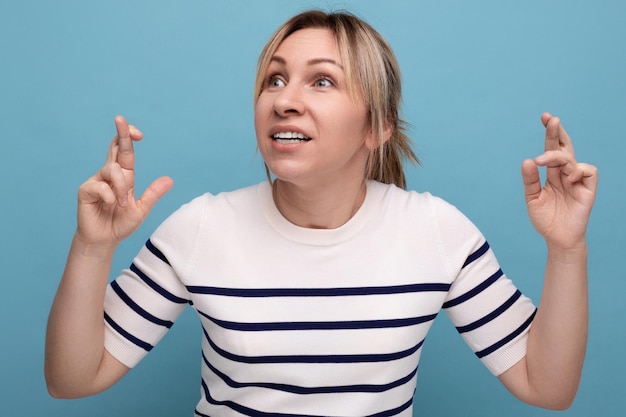 Closeup photo of a blond young woman crossing her fingers and praying on a blue background with