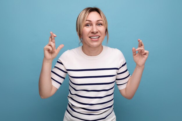 Closeup photo of a blond young woman crossing her fingers and praying on a blue background with