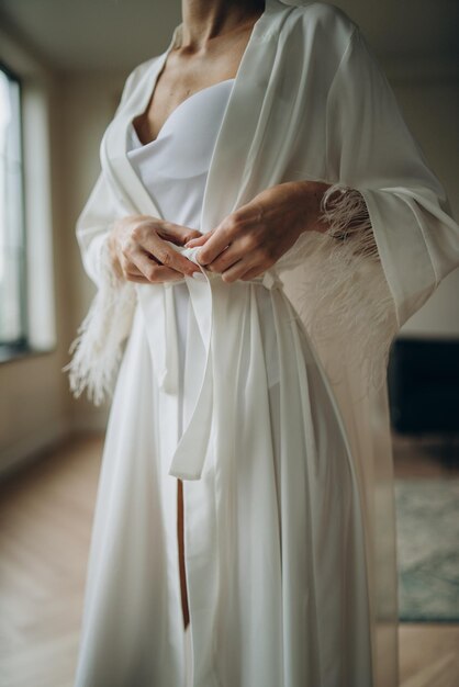 Closeup photo of a bride in silk dressing gown holding wedding dress infront of the window Wedding morning bride's preparations