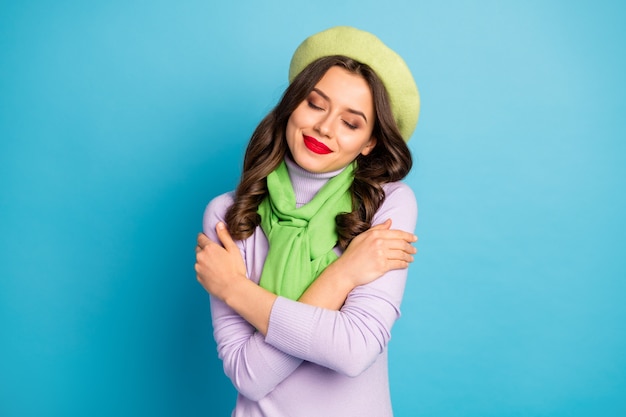 Closeup photo of cute lady red lips eyes closed hugging herself peace inner harmony concept wear green beret hat purple turtleneck scarf isolated blue color wall