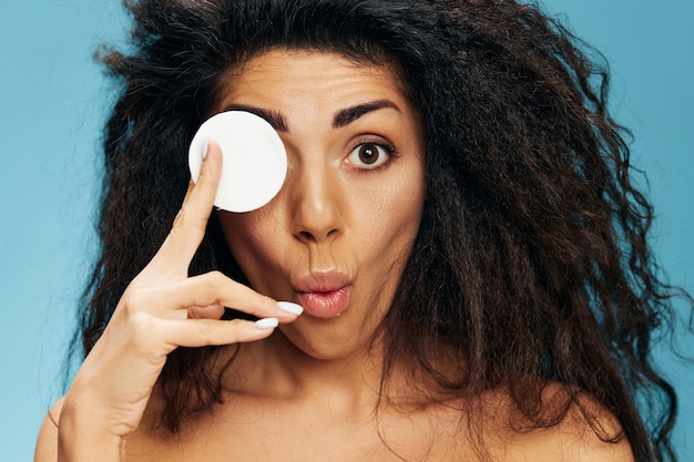 Closeup photo of excited pretty naked young latin curly woman\
posing with cotton pads covering her eye and looking at camera\
using makeup removing beauty products isolated blue background