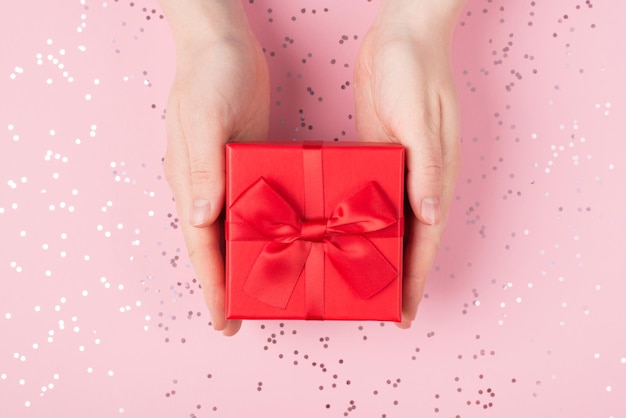 Primo piano di mani femminili che danno una confezione regalo rossa su uno sfondo luccicante di colore pastello