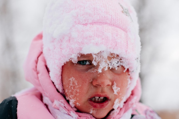 雪に覆われた悲しい顔をした女性の子供のクローズアップ写真と、森の中でピンクの冬の服を着て泣きそうになっている白い色と雪でいっぱいの驚くべき背景