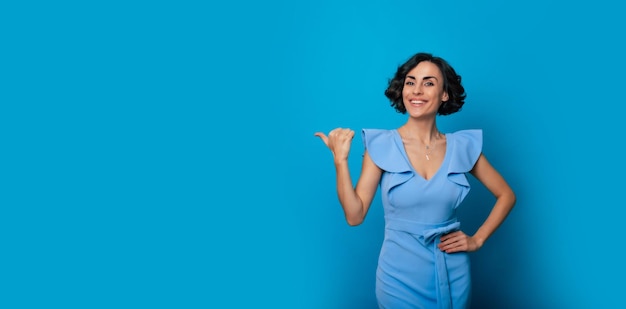 Closeup photo of a happy beautiful young woman in a blue dress who is pointing with help of her finger and isolated on blue background