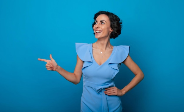 Closeup photo of a happy beautiful young woman in a blue dress who is pointing with her index finger and isolated on blue background