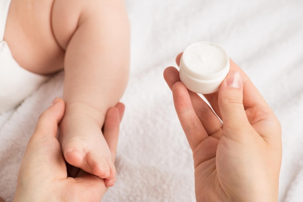 Photo closeup photo of mother's hands holding newborn's leg and little round cream bottle on isolated white blanket background