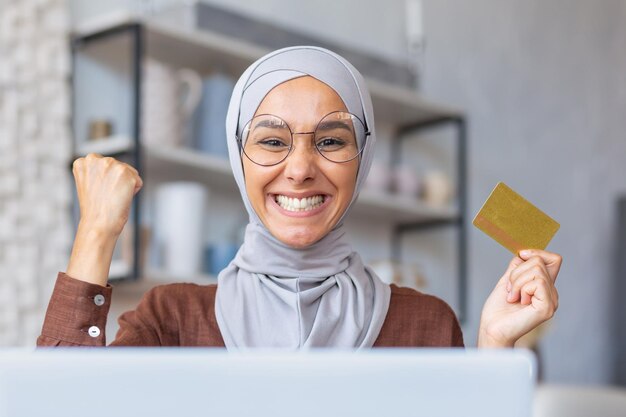 ラップトップを使用して、幸せなクレジット カードを保持している自宅で座っているヒジャーブのイスラム教徒の若い女性のクローズ アップ写真