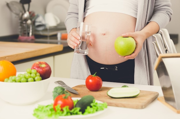 Closeup photo of pregnant woman posing on kitchen with apple and glass of water