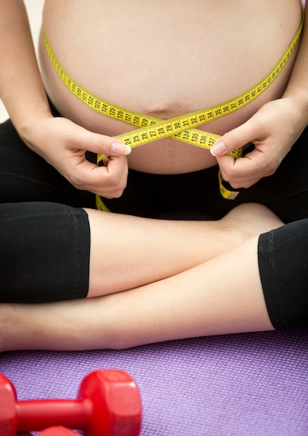 Closeup photo of pregnant woman sitting on fitness mat and measuring belly with tape