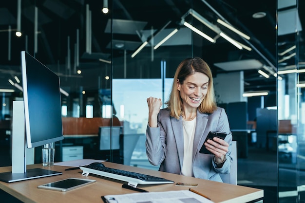 Closeup photo of a successful business woman working in a modern office happy boss rejoices in the good news looks at the phone and celebrates success and victory