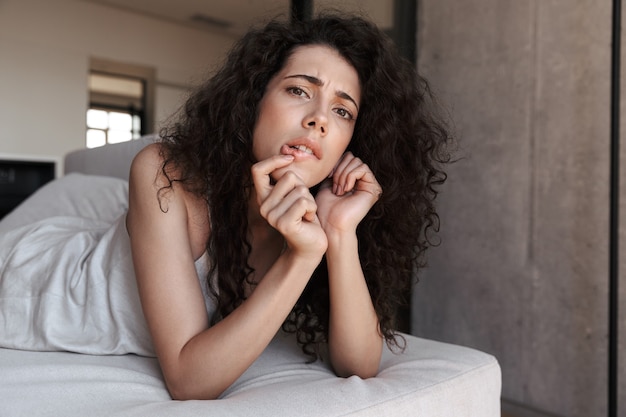 Closeup photo of uptight thoughtful woman with long curly hair wearing silk leisure clothing lying in hotel bed or sofa, and biting lips while propping up her head with hand