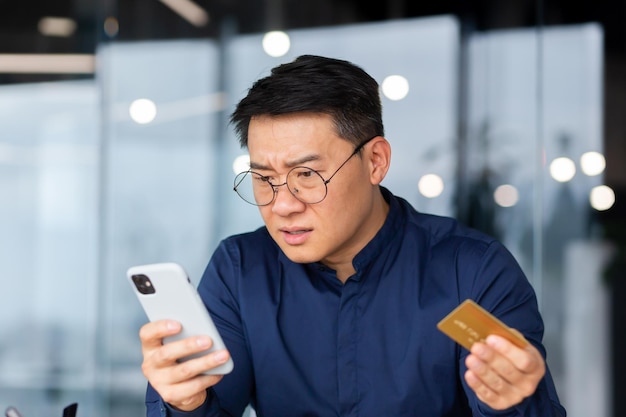 Closeup photo a worried young asian man in glasses holds a credit card and look shocked at the phone