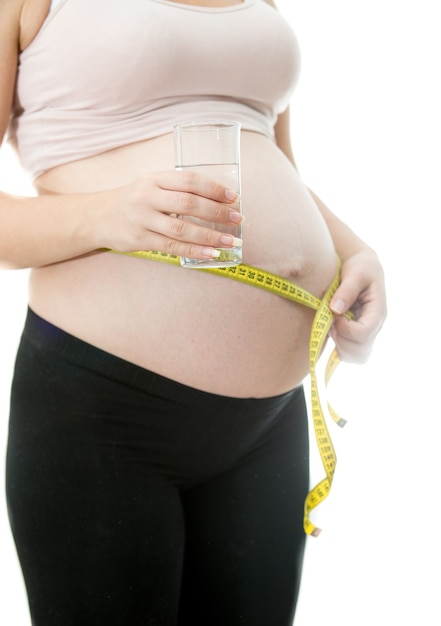 Closeup photo of young pregnant woman holding glass of water and measuring belly.