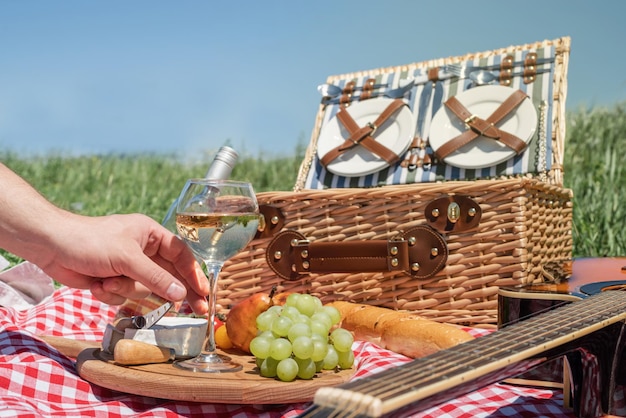 ワイングラスを持っている草の男性の手に飲み物と食べ物とピクニックバスケットのクローズアップ