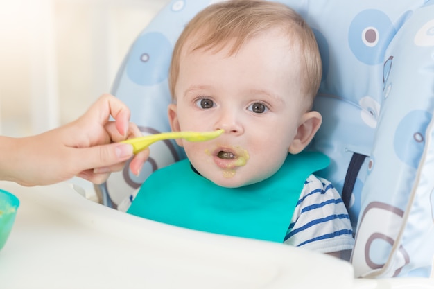 スプーンからフルーツソースを食べるエプロンの愛らしい赤ちゃんのクローズアップの肖像画