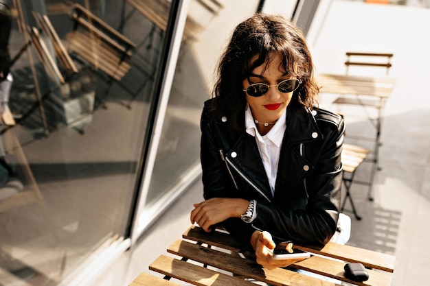 Photo closeup portrait of adorable brunette girl in black jacket is drinking coffee on summer terrace in sunshine lovable lady sitting near window and chatting online with friends