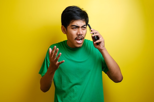 Closeup portrait angry young asian man, guy mad student, pissed off employee shouting while on phone isolated yellow wall background. Negative human emotion face expression feeling attitude