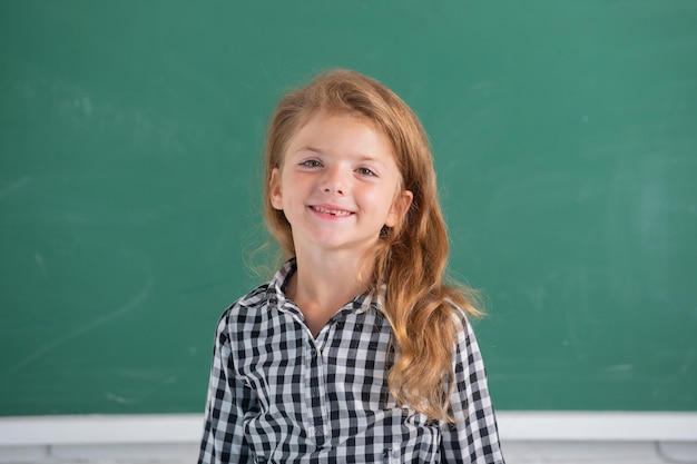 Крупным планом портрет привлекательной маленькой веселой девочки, сидящей на рабочем столе в классе