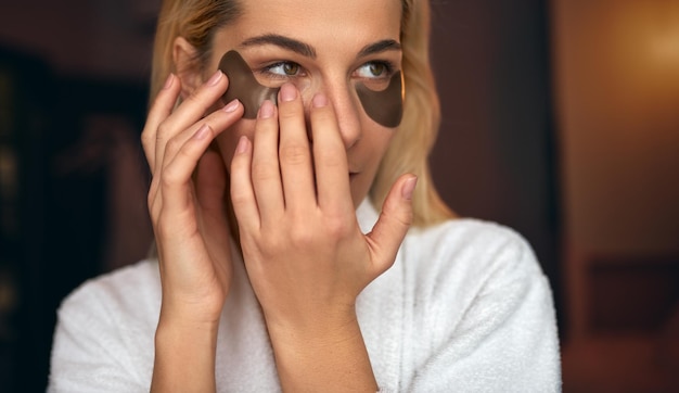 Photo closeup portrait of attractive young woman with natural makeup puts patches under the eyes from wrinkles and dark circles beauty spa at home attractive young woman in bathrobe spending time at home